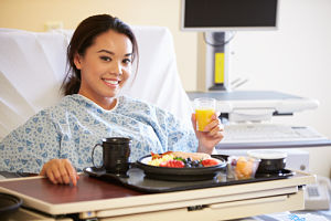Female patient is sitting up in a swing bed, with a plate of breakfast in front of her, on a rolling tray table, and a glass of orange juice in one hand